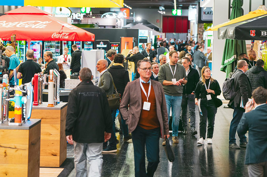 Belebte Messehalle mit zahlreichen Besuchern, Ausstellerständen und Getränkemarken im Hintergrund.