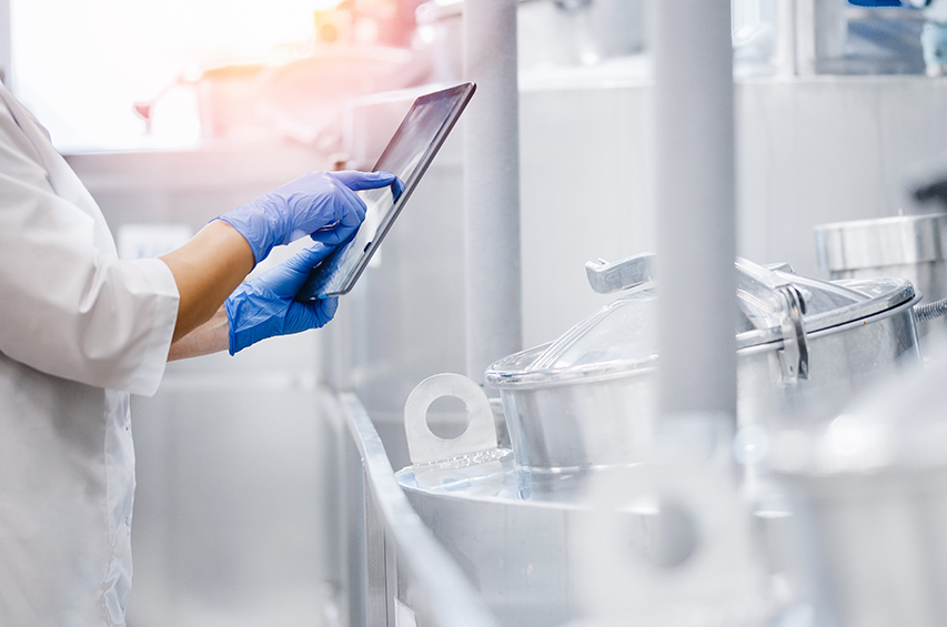 Person in Schutzkleidung überwacht mit einem Tablet die Lebensmittelproduktion an Edelstahltanks
