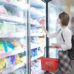 Eine Kundin wählt in einem Supermarkt ein Produkt aus einem geöffneten Tiefkühlschrank. In der Hand hält sie einen roten Einkaufskorb.