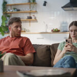 Mann und Frau sitzen auf einem Sofa in einer Wohnküche, beide schauen auf ihr Smartphone, der Mann blickt skeptisch zur Seite.
