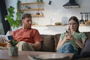 Mann und Frau sitzen auf einem Sofa in einer Wohnküche, beide schauen auf ihr Smartphone, der Mann blickt skeptisch zur Seite.