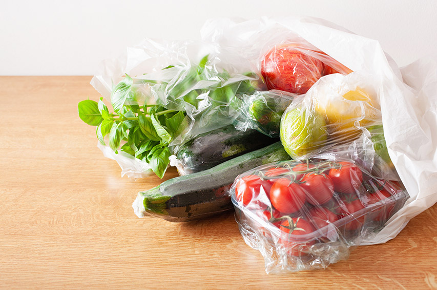 Frisches Gemüse und Obst in Kunststoffverpackungen auf einem Holztisch: Zucchini, Basilikum, Kirschtomaten, Apfel und Zitrone.