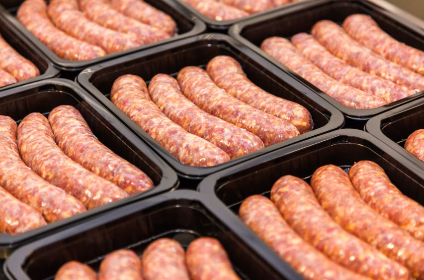Mehrere rohe Fleischwürste liegen dicht nebeneinander in einer Verpackungsschale. Das Bild zeigt unverarbeitete Würstchen, die für die weitere Verarbeitung oder Verpackung in der Fleischwarenindustrie vorbereitet sind.