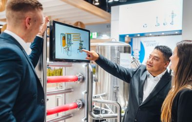 Ein Aussteller erklärt zwei Besuchern an einem Bildschirm eine Brauanlage, im Hintergrund Edelstahltanks und Messestand.