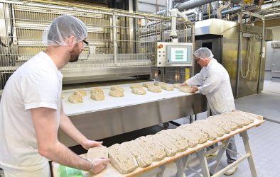 Mitarbeiter in einer Bäckerei bereiten Brotlaibe auf einem Förderband vor; im Hintergrund moderne Backtechnik.