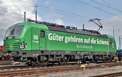 Grüne Elektro-Lokomotive der DB Cargo mit der Aufschrift 'Güter gehören auf die Schiene' auf einem Bahngleis. Die Oberleitung ist angeschlossen, im Hintergrund sind weitere Schienen und Strommasten zu sehen.
