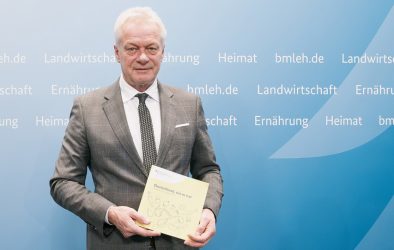 Bundesernährungsminister Alois Rainer hält den Ernährungsreport 2025 vor einer blauen Wand mit Ministeriumsbegriffen hoch.