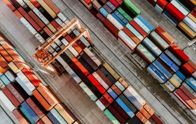 Luftaufnahme eines Containerhafens mit farbenfrohen, gestapelten Frachtcontainern und einem großen Verladekran. Die Container sind in langen Reihen auf dem Betonboden angeordnet, bereit zum Transport oder zur Weiterverladung.