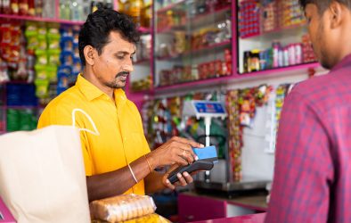 Ladenbesitzer in einem indischen Kirana-Shop verarbeitet eine Kartenzahlung, im Hintergrund vollbepackte Regale mit Lebensmitteln und Snacks