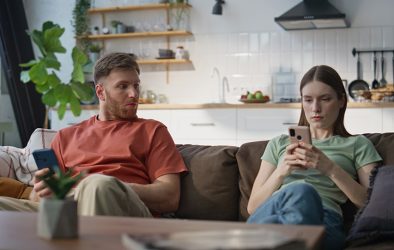 Mann und Frau sitzen auf einem Sofa in einer Wohnküche, beide schauen auf ihr Smartphone, der Mann blickt skeptisch zur Seite.