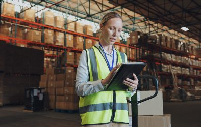Mitarbeiterin in einer Lagerhalle prüft Liefer- und Bestandsdaten auf einem Tablet zwischen Hochregalen mit Waren.