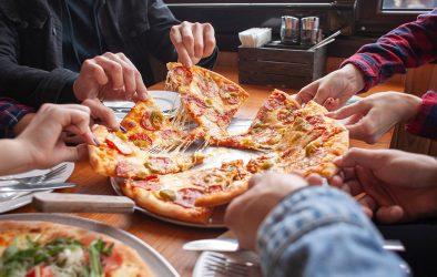 Mehrere Personen greifen gleichzeitig nach Stücken einer frisch gebackenen Pizza mit Salami, Käse und Peperoni.