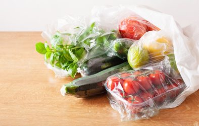 Frisches Gemüse und Obst in Kunststoffverpackungen auf einem Holztisch: Zucchini, Basilikum, Kirschtomaten, Apfel und Zitrone.