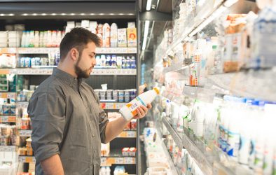 Ein Mann steht im Supermarkt vor einem Kühlregal und prüft aufmerksam das Etikett einer Milchflasche – Symbolbild für bewussten Einkauf und Preisvergleich.