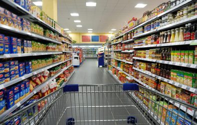 Blick durch einen Supermarktgang aus der Perspektive eines Einkaufswagens, beidseitig dicht gefüllte Regale mit Lebensmitteln