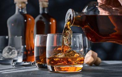 Whiskey wird aus einer Karaffe in ein Glas eingeschenkt, im Hintergrund stehen weitere Flaschen und Gläser.