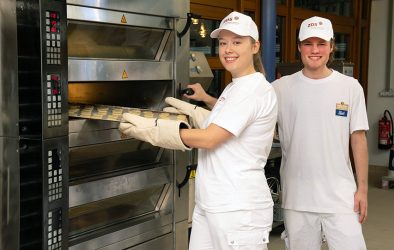 Zwei Auszubildende in weißer Arbeitskleidung stehen in einer Backstube. Eine Person schiebt mit hitzebeständigen Handschuhen ein Blech in einen industriellen Etagenbackofen, eine zweite Person steht daneben.